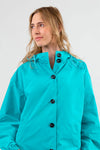 Someone wearing the looselyboho ELECTRIC CYAN WINDBREAKER, featuring large black buttons and a high collar, glances to the side. The lightweight, water-resistant fabric of the windbreaker contrasts with their long, wavy blonde hair against a plain light gray background.