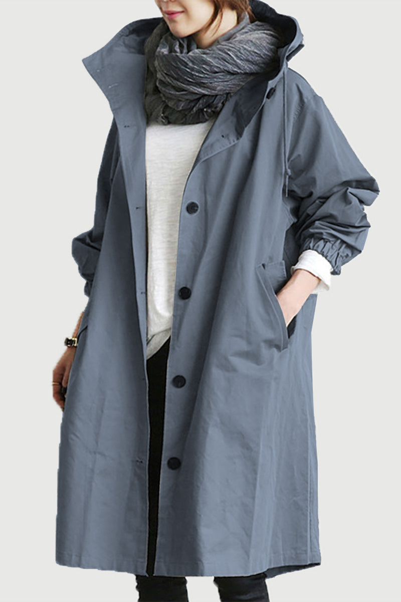 A person is wearing The WJacket's Water Resistant Oversized Hooded Windbreaker Rain Jacket in gray, embodying a timeless style with black buttons. They have their hands in the jacket pockets and are also wrapped in a thick gray scarf around their neck. The jacket, with its wide collar, is paired casually with a white long-sleeve shirt underneath.