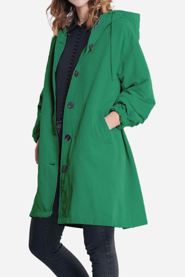 A person wearing The WJacket's Water Resistant Oversized Hooded Windbreaker Rain Jacket in green, featuring large black buttons, is standing with one hand in a pocket. This timeless style is paired with black pants and a black top. The person's face is not visible in the image.