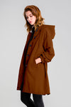 A person with shoulder-length hair wears the "Burnt Brown Windbreaker" by looselyboho, featuring a rust color, large black buttons, and an adjustable hood made from lightweight, water-resistant fabric. They stand with hands in pockets against a gray background, gazing downward.