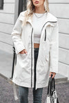 A person with long blonde hair is wearing a BELTED FROST WHITE WINDBREAKER by looselyboho over a white cropped top and dark jeans. They are holding a black and white handbag and standing on a city street with gray buildings in the background.