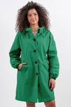 A person with curly hair wears an EMERALD GREEN WINDBREAKER by The Windbreaker Jacket made from water-resistant material, complete with black buttons and adjustable hood. They have their left hand in their pocket and are standing against a plain white background.