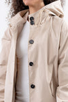 A person with curly hair is wearing a light beige, lightweight water-resistant jacket (BISCUIT BEIGE WINDBREAKER by looselyboho) with black buttons over a white top. The stylish rain-jacket features an adjustable hood and drawstrings. They are standing against a plain background, with only the upper torso visible.