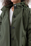 A person wearing The Windbreaker Jacket's OLIVE GREEN WINDBREAKER, featuring large black buttons down the front. The coat has a high collar, and the adjustable hood is cinched with a drawstring. The person sports a white top underneath, and only the lower part of their face is visible.